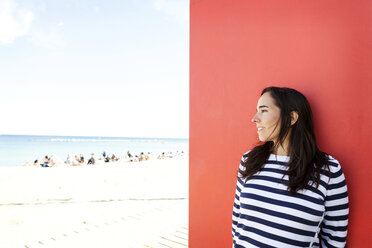 Spanien, Barcelona, Frau in gestreiftem Pullover lehnt an roter Wand und schaut zum Strand - VABF000449