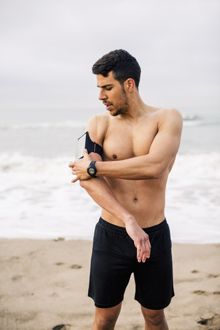 Sportlicher junger Mann am Strand beim Einstellen des Handys, lizenzfreies Stockfoto