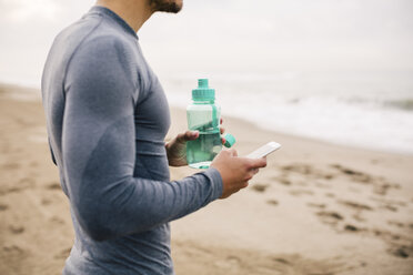 Sportlicher junger Mann mit Handy und Trinkflasche am Strand - EBSF001317