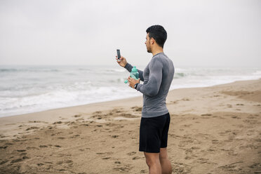 Sportlicher junger Mann mit Handy und Trinkflasche am Strand - EBSF001318