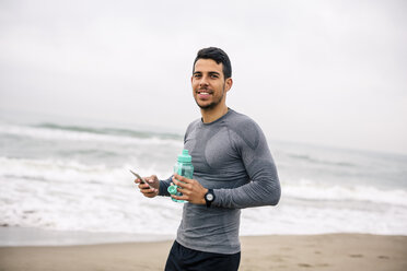 Lächelnde Sportlerin mit Handy und Trinkflasche am Strand - EBSF001319