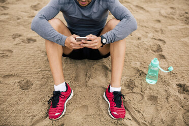 Sportlicher junger Mann mit Handy und Trinkflasche am Strand - EBSF001321