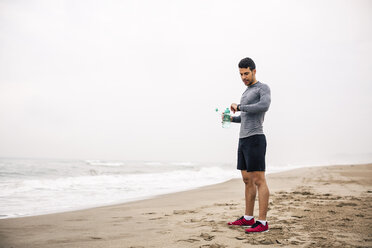 Sportive young man with drinking bottle on the beach looking at watch - EBSF001322