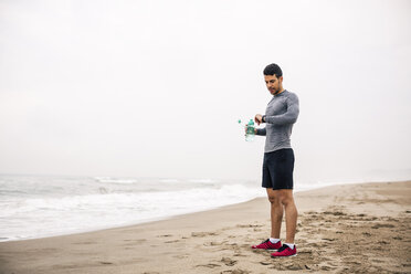 Sportlicher junger Mann mit Trinkflasche am Strand und Blick auf die Uhr - EBSF001322