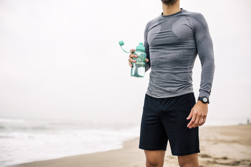 Sportlicher junger Mann mit Trinkflasche am Strand stehend - EBSF001323