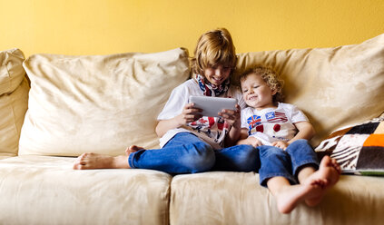Zwei lächelnde Jungen sitzen auf einer Couch und benutzen ein digitales Tablet - MGOF001741