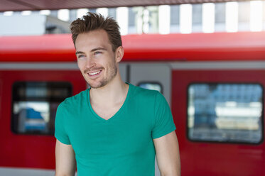 Porträt eines lächelnden jungen Mannes mit Bartstoppeln auf einem Bahnsteig - DIGF000320