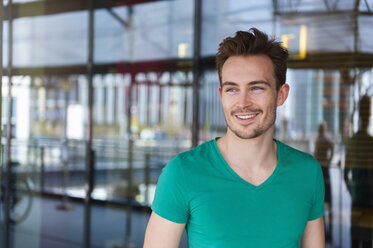 Portrait of smiling young man with stubble standing in front of glass facade - DIGF000322