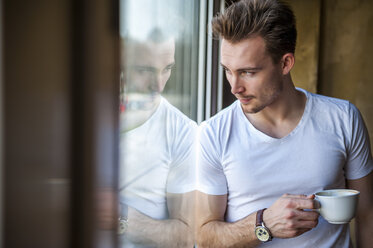 Junger Mann mit Tasse Kaffee schaut durch das Fenster - DIGF000343