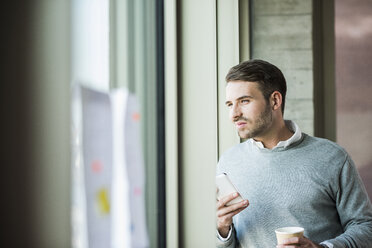 Junger Mann mit Mobiltelefon schaut aus dem Fenster - UUF007032