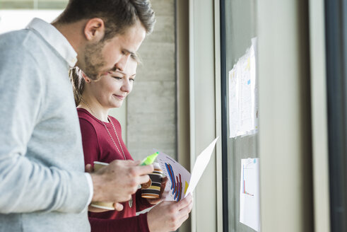 Zwei Kollegen am Bürofenster, die auf Papier schauen - UUF007042