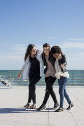 Three young women walking arm in arm by the sea - MAUF000447
