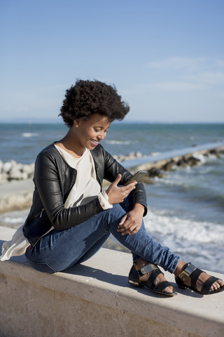 Junge Frau sitzt am Meer und benutzt ihr Smartphone, lizenzfreies Stockfoto