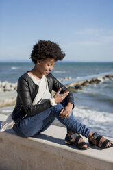 Young woman sitting by the sea, using smart phone - MAUF000450