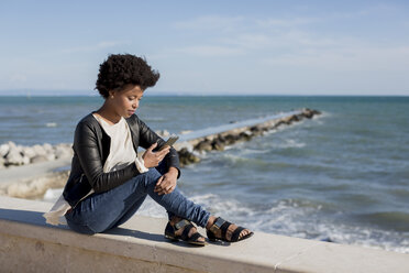 Young woman sitting by the sea, using smart phone - MAUF000451