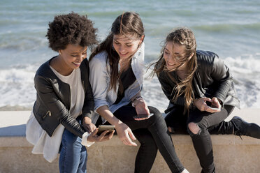 Drei junge Frauen sitzen an der Wand und schauen auf ihr Smartphone - MAUF000452