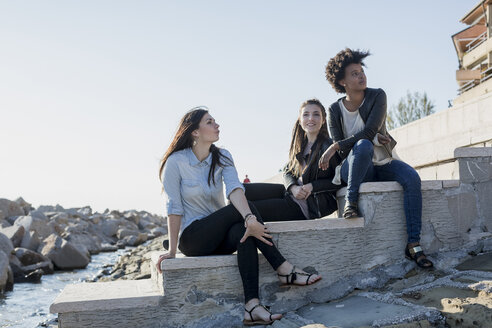 Drei junge Frauen sitzen auf einer Mauer am Meer - MAUF000482