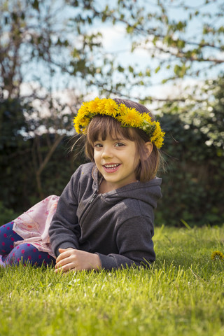 Porträt eines lächelnden kleinen Mädchens, das auf einer Wiese liegt und einen Blumenkranz aus Löwenzahn trägt, lizenzfreies Stockfoto