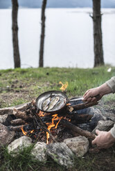 Man frying rudd at camp fire at shore of a lake, partial view - DEGF000787