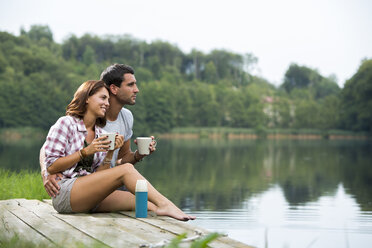 Entspanntes junges Paar mit Kaffeetassen, das auf einem Steg am See sitzt und in die Ferne schaut - ZOCF000052