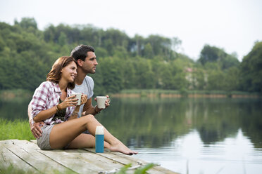 Relaxed young couple with coffee mugs sitting on a jetty at lake looking at distance - ZOCF000052