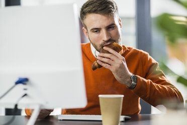 Junger Mann mit einem Snack am Schreibtisch im Büro - UUF007103