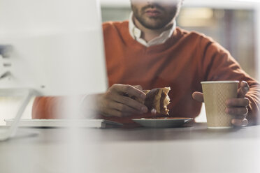 Junger Mann mit einem Snack am Schreibtisch im Büro - UUF007105