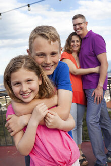 Porträt eines Jungen, der seine kleine Schwester umarmt, während die Eltern im Hintergrund stehen - VABF000470