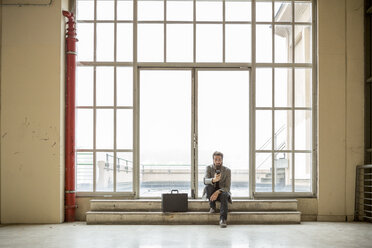 Hipster-Geschäftsmann mit Aktentasche wartet vor einer Tür in einem Industriegebäude - ZOCF000094