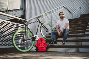 Lächelnder junger Mann sitzt auf einer Treppe neben einem Fixie-Fahrrad - RAEF001124