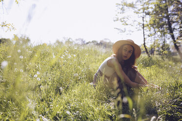 Young woman wearing straw hat sitting on a meadow - GIOF000913