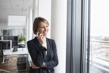 Lächelnde Geschäftsfrau mit Mobiltelefon am Fenster - RBF004474