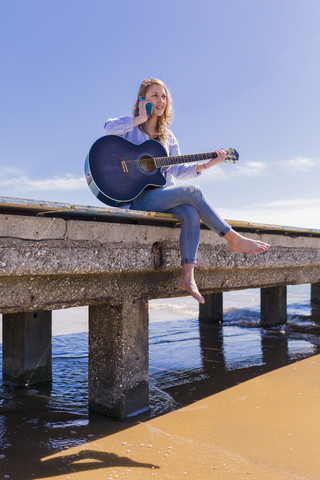 Frau mit Smartphone und Gitarre auf dem Bootssteg sitzend, lizenzfreies Stockfoto