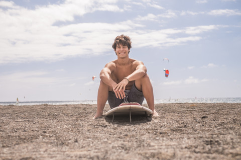 Porträt eines lächelnden jungen Mannes, der auf seinem Surfbrett am Strand sitzt, lizenzfreies Stockfoto