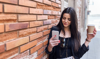 Junge schöne Frau, die einen Kaffee zum Mitnehmen und ein Smartphone trinkt und sich an die Wand lehnt - MGOF001808