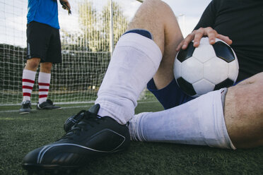 Beine eines Mannes mit Fußballkleidung, der mit einem Ball auf dem Rasen sitzt - ABZF000459