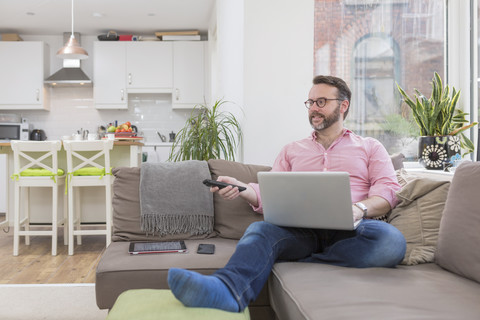 Älterer Mann sitzt auf der Couch und benutzt Laptop und Fernbedienung, lizenzfreies Stockfoto