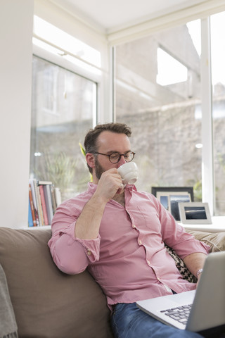 Älterer Mann auf der Couch, der Kaffee trinkt und einen Laptop benutzt, lizenzfreies Stockfoto