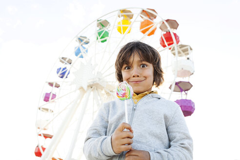 Porträt eines kleinen Jungen mit Lutscher, der vor einem Riesenrad lustige Gesichter zieht - VABF000481