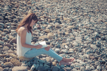 Frau sitzt am steinigen Strand und schreibt in ihr Notizbuch - SIPF000417