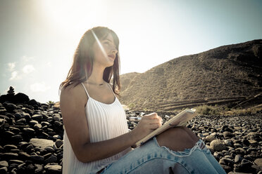 Woman with notebook sitting on stony beach at backlight - SIPF000422