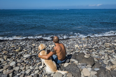 Rückenansicht eines Mannes, der mit seinem Hund am steinigen Strand sitzt und auf das Meer schaut - SIPF000424
