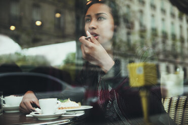 Young woman sitting behind windowpane of a cafe eating cake - ZEDF000091