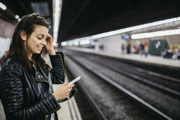 Lächelnde junge Frau auf dem Bahnhof, die auf ihr Handy schaut - JRFF000626