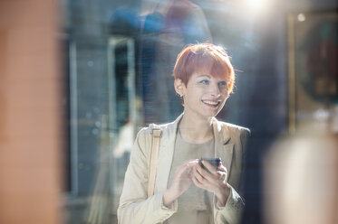 Lächelnde junge Frau mit Handy in der Hand betrachtet ein Schaufenster - DIGF000395