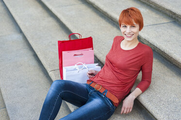Lächelnde junge Frau auf der Treppe sitzend mit Einkaufstüten neben sich - DIGF000419