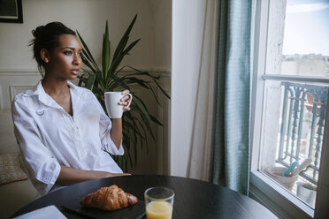 Junge Frau sitzt beim Frühstück mit einer Tasse Kaffee und schaut durch das Fenster - ZEDF000096