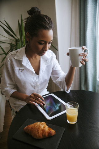 Junge Frau verwendet digitales Tablet am Frühstückstisch, lizenzfreies Stockfoto
