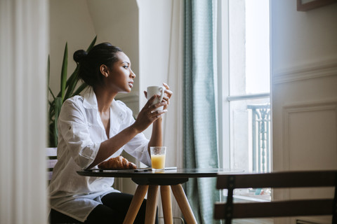 Junge Frau sitzt beim Frühstück mit einer Tasse Kaffee und schaut durch das Fenster, lizenzfreies Stockfoto