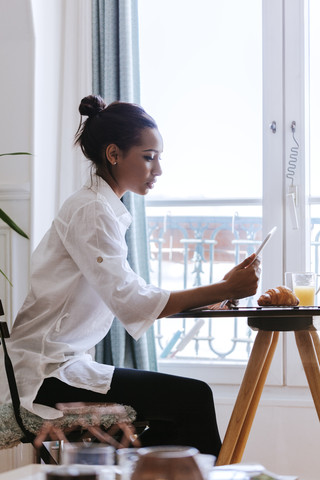 Junge Frau verwendet digitales Tablet am Frühstückstisch, lizenzfreies Stockfoto